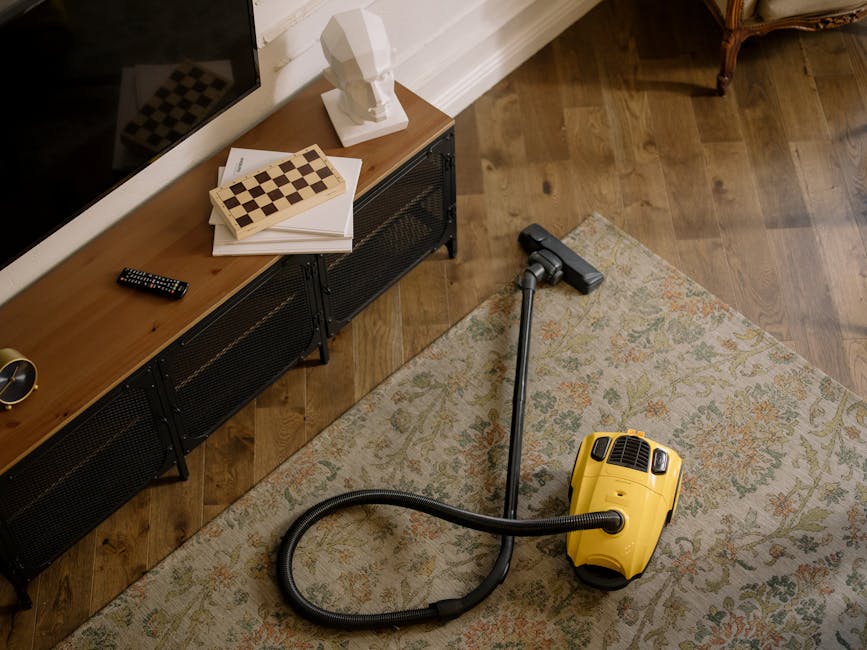 A person kneeling on a patterned area rug in a residential room, using a yellow and black vacuum cleaner for surface cleaning and deep cleaning. The individual is focused on the vacuum's inlet, which 