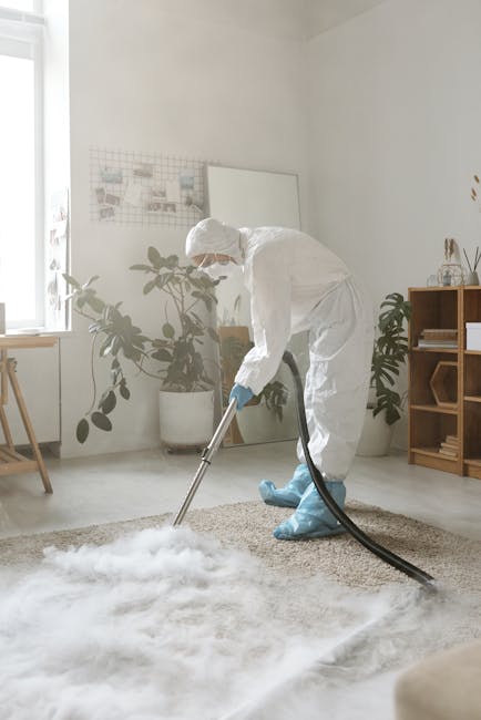 Close-up of a wet vacuum cleaner with a clear, water-filled container and a black base, connected to a flexible, patterned hose, cleaning a light pink carpet in an indoor setting with a plain white wa